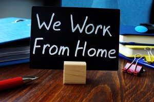 A small blackboard sign with the handwritten message "We Work From Home" is placed on a wooden desk. The background includes a blue binder, notebooks, a red pen, a few paper clips, and other office supplies—a perfect setup for anyone exploring careers in government.