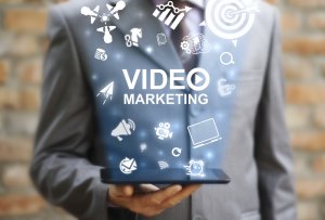A person in a suit holds a tablet displaying icons related to digital marketing, including graphs, a bullseye, a megaphone, and computer monitors. The central text reads "VIDEO MARKETING," while the background features a blurred brick wall — ideal for those pursuing government jobs to enhance their communication strategies.