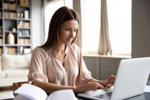 A person with long brown hair wearing a light pink blouse is sitting at a desk, working on a laptop, researching careers in government. There is a bookshelf in the background with books and decorative items. The room is well-lit with natural light coming through large windows.
