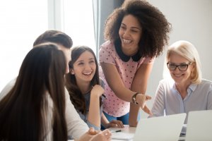 A leader among the group of diverse colleagues is standing and gesturing energetically while engaging in a lively discussion around a table with laptops. The others sit and smile, reflecting a friendly and collaborative work environment.