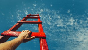 A close-up of a hand grasping a red ladder, with the ladder extending upwards into a blue sky with scattered clouds. The perspective gives a sense of climbing towards the sky, evoking a feeling of power and determination.