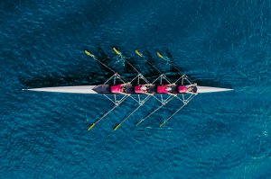 An overhead view of a four-person rowing team in a sleek, white rowing shell cuts through deep blue water. The rowers, dressed in red attire and synchronized through flawless teamwork, each hold yellow oars, creating a dynamic pattern in the water.