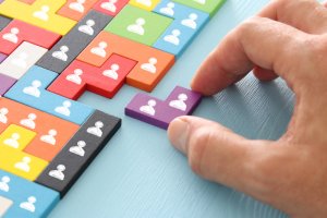 A hand is placing a purple puzzle piece with a white person icon among several other colorful puzzle pieces, each symbolizing leaders. The pieces are arranged on a light blue surface.