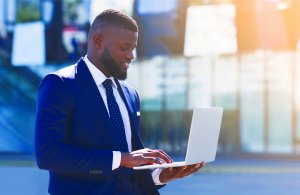 A man in a blue suit stands outdoors, using a laptop to update his resume. The background is blurred, showing a modern, urban environment with glass buildings. Sunlight streams from the top right corner, creating a warm glow on the scene.