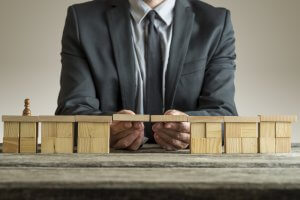 A person in a business suit places a wooden bridge plank between two sets of wooden blocks on a table, connecting them. A small pawn stands on one block set, symbolizing crossing or transition, much like starting a new chapter in one’s resume. The background is plain and neutral.