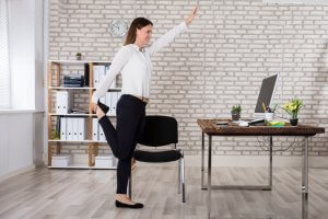 A woman in office attire stands beside her desk, performing a stretching exercise by holding one foot behind her with one hand and extending the other hand forward. In the background, there is a brick wall, shelves with binders, and a clock.