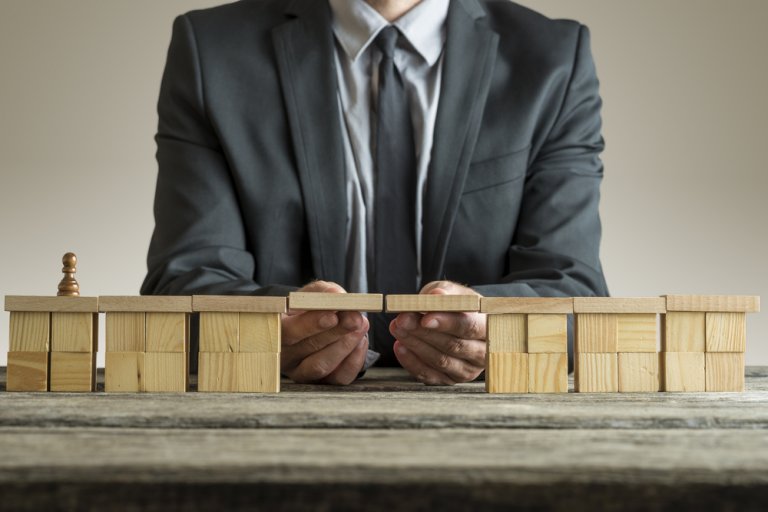 A person in a business suit places a wooden bridge plank between two sets of wooden blocks on a table, connecting them. A small pawn stands on one block set, symbolizing crossing or transition, much like starting a new chapter in one’s resume. The background is plain and neutral.