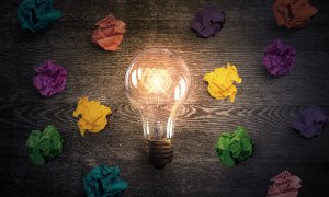 A glowing light bulb lies on a dark wooden surface surrounded by crumpled pieces of colorful paper, including purple, yellow, orange, and green. The light from the bulb casts a warm glow, as if leading the vibrant colors of the paper into vivid relief.