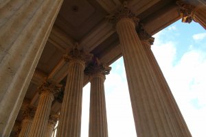 Low-angle view of several tall, ornate classical columns supporting a roof, with a bright sky in the background. Amidst the grandeur, it's easy to forget these structures have stood firm through countless challenges, from wars to pandemics like Covid-19. The intricate capitals at the top add to the architectural detailing.