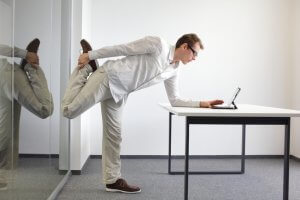 A man wearing a white shirt and beige pants is stretching his leg by pulling his foot towards his back while leaning forward on a white desk. He is looking at a tablet placed on the desk in a modern, minimalistic office setting, seamlessly incorporating exercise into his workday routine.