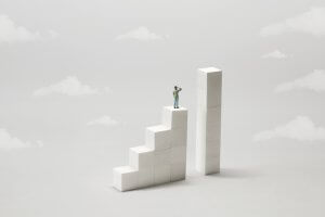 A small figurine of a person stands atop a staircase of four ascending white blocks, much like building a resume step by step, looking up at a taller adjacent column composed of five stacked blocks. The background is a light grey with subtle, fluffy clouds.