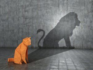 An orange origami cat sits on a gray tiled floor, casting a shadow on a concrete wall. The shadow, resembling a lion, signifies a powerful transformation theme. The setting is minimalistic, with a focus on the imaginative and the bold contrast.
