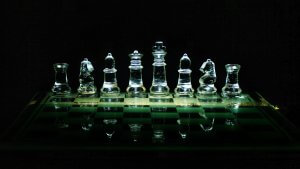 A row of transparent glass chess pieces, including a rook, knight, bishop, queen, king—the leader—bishop, knight, and rook, are arranged on a glass chessboard. The set is illuminated dramatically against a dark background.