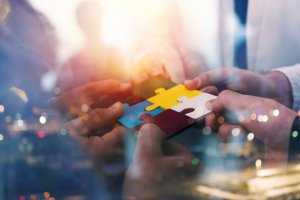 A group of people collaboratively fitting colorful puzzle pieces together, symbolizing effective teamwork. The background features a soft-focus cityscape with glowing lights, enhancing the sense of unity and cooperative effort.