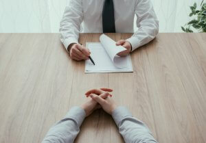 Two individuals are sitting across from each other at a wooden table. One person, visible from the waist down, holds a pen and a resume. The other person, visible only by their clasped hands, appears to be in discussion.
