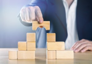 A person in a business suit is placing a wooden bridge piece between two stacks of wooden blocks, symbolizing connection and building. The surface is light-colored, and the background is slightly blurred, emphasizing the focus on the hand, blocks, and metaphorical steps to enhance one's resume.