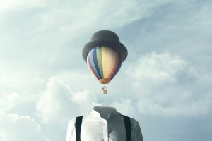A surreal image of a headless person wearing a white shirt, black suspenders, and a grey bowler hat. A colorful hot air balloon sits in place of the head, challenging our assumptions, with an expansive cloudy sky in the background.