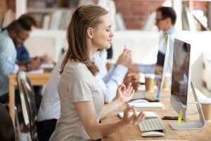 A woman sits at a desk in front of a computer, eyes closed and hands in a meditative pose. She appears calm and relaxed, as if finishing a mental exercise. Other people are working at desks in the background, focused on their tasks. The workspace is bright and modern.