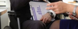 A person with cerebral palsy in a wheelchair holds a clipboard with a statistical report, while another person sitting across gestures with a pen. Both are engaged in a discussion and wearing formal attire.