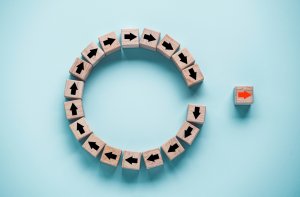 Wooden blocks with black arrows arranged in a circular pattern on a light blue background. One block with an orange arrow, symbolizing leaders taking a different direction, is positioned outside the circle, suggesting breaking away from the norm.