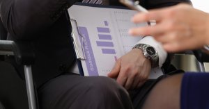 A person dressed in a suit holds a clipboard with a document titled "Statistics Report" featuring bar graphs. Another person's hand, possibly of someone with cerebral palsy, is visible in the foreground, holding a smartphone. The focus remains on the clipboard and the hands.