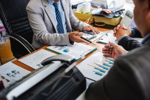 Three business professionals in formal attire are engaged in a meeting around a table. The table is covered with documents and charts displaying financial data and employee performance metrics. One person is holding and pointing with a pen, discussing the presented figures. Various office supplies are in the background.