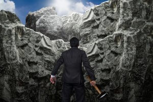 A man in a dark suit stands at the base of a steep, rocky cliff, holding a sledgehammer. He looks up toward the top of the cliff, where sunlight is breaking through the clouds, illuminating the rocky terrain—much like leaders seeking to illuminate new paths.