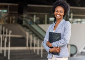 A woman in a light blue blazer is smiling while holding a black folder, likely containing her resume. She is standing outdoors, with modern building features and an escalator in the background.