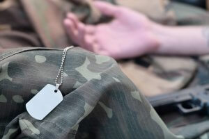 Close-up of military dog tags resting on camouflage fabric. In the background, a person's outstretched arm is visible, hinting at them lying down. The image emphasizes military experience, with the focus on the dog tags in the foreground.