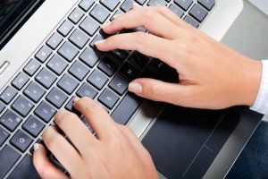 Close-up image of two hands typing on a silver laptop keyboard. The fingers are positioned on the keys, with both hands actively engaged despite visible signs of cerebral palsy. The laptop's touchpad is visible below the keyboard, and part of a blue shirt sleeve is noticeable on one wrist.