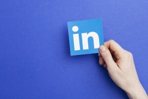 A hand holds a blue square card with the white letters "in" on it, symbolizing the LinkedIn logo, against a plain blue background, perfectly capturing the essence of LinkedIn's clean and professional branding.