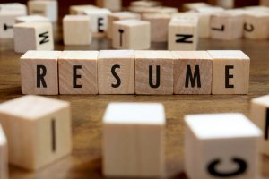 Wooden blocks spelling out the word "RESUME" are prominently arranged in the center of the image, with scattered blocks bearing random letters in the background on a rustic wooden surface.