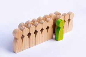 A group of light-colored wooden human-shaped figures stand in a row. One green wooden figure, symbolizing career leadership, stands facing the group, positioned as if leading or confronting them. The background is white and plain.