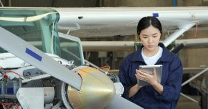 A person with military experience wearing a blue jumpsuit stands next to an aircraft engine inside a hangar, holding a tablet. With part of the propeller and engine visible, the individual appears focused, likely performing maintenance or inspections.