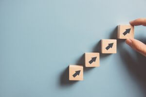 A hand places the fourth wooden cube with a black arrow on it, completing a diagonal sequence of four ascending cubes, symbolizing employee performance. The background is a solid light blue, suggesting a concept of growth or progress.