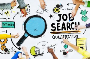 Top view of a table with a colorful job search concept illustrated. Hands of diverse individuals hold pens and markers, collaborating on the design. Elements include a magnifying glass, words "Job Search," "Qualification," "Resume," "Interview," and various job-related icons.