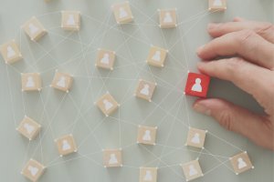 A hand is holding a red wooden block with a white person icon on it, symbolizing a manager. The block is placed among several other beige blocks with similar icons, all connected by white lines and dots forming a network on a light green background.