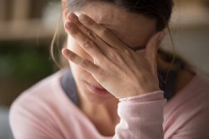 A close-up of a person in a pink long-sleeve shirt covering their face with their hand, conveying a sense of anxiety and frustration. The background is blurred, emphasizing their emotional expression.