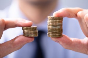 Close-up of a person holding two stacks of coins, highlighting the issue of pay equity. The left hand grasps a small stack, while the right hand holds a noticeably larger one. The background is blurred, drawing attention to the disparity between the coins and hands.