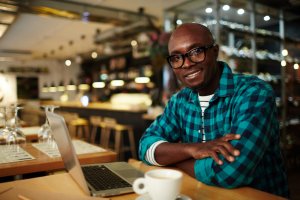 A man wearing glasses and a plaid shirt sits at a wooden table in a cozy café, smiling at the camera. He has a laptop open in front of him and a coffee cup nearby, possibly working on job tasks. The background features barstools, hanging lights, and a modern, inviting atmosphere.