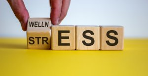 A hand turns a wooden block to change the word "STRESS" into "WELLNESS." The blocks rest on a yellow surface, with "WELLNESS" partially visible. This reflects the concept of transforming anxiety and stress into well-being.