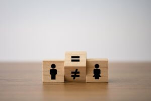 Three wooden blocks on a table: the left block has a male symbol, the right block has a female symbol, and the middle block features an equals sign on top and a not equal sign facing the front, highlighting the discussion on gender equality and pay equity.