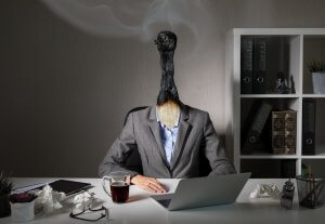 A person in a suit sits at a desk with a burnt matchstick instead of a head, symbolizing burnout. The desk features a laptop, crumpled paper, coffee, glasses, and office supplies. In the background, there are shelves with books on medical care, binders, and a small plant.