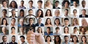 A mosaic of 50 diverse people smiling, arranged in a grid pattern. Central to the image, someone holds a magnifying glass that highlights one person's face, slightly enlarging it as if they are up for an important job. Everyone is against neutral backdrops, looking directly at the camera.