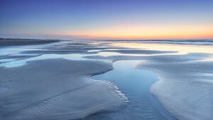 A serene beach at twilight with shallow tidal pools reflecting the colorful sky above. The horizon is illuminated with soft hues of orange and purple as the sun sets, casting a tranquil glow over the smooth, wet sand and gentle waves of the ocean, managing to create a perfect moment of peace.