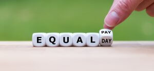 A hand is shown arranging white dice blocks that spell out "EQUAL PAY DAY" on a wooden surface, highlighting the importance of pay equity. The background is green and slightly blurred.