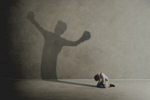 A person dressed in a white shirt and suspenders kneels on the ground with blue boxing gloves, head bowed. On the plain wall behind, their shadow is cast in a triumphant victory pose with arms raised and fists clenched, wearing the same boxing gloves—symbolizing true winners.
