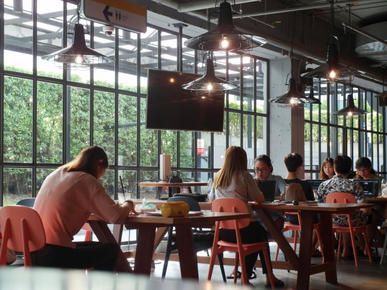A modern, well-lit coworking space with large windows revealing greenery outside. Several people sit at wooden tables, working on laptops or reading. Pendant lights hang from the ceiling, illuminating the flexible work environment.