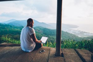 A person with a beard and a white shirt is sitting on a wooden platform overlooking a scenic mountain landscape. They are using a laptop resting on their knees, connected to the local wifi, and a smartphone lies beside them. The sky is partly cloudy, and there is a town in the distance.