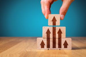 A hand is stacking wooden blocks with arrows pointing upward. The blocks are arranged in a pyramid shape on a wooden surface against a blue background, symbolizing more effective growth or progress.